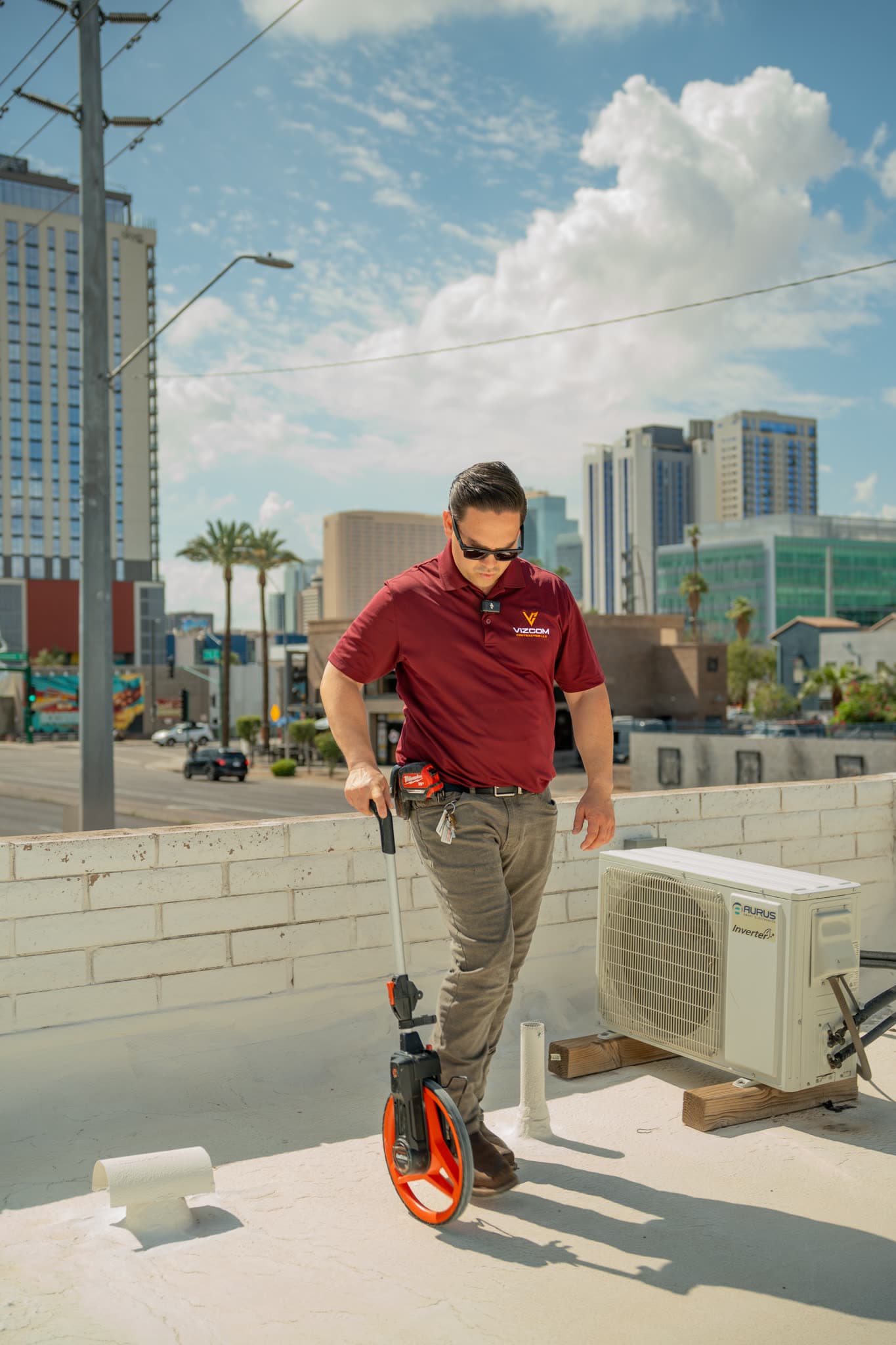 Omar Vizcarra measuring a white commercial flat roof with a wheel, downtown high rises behind him.
