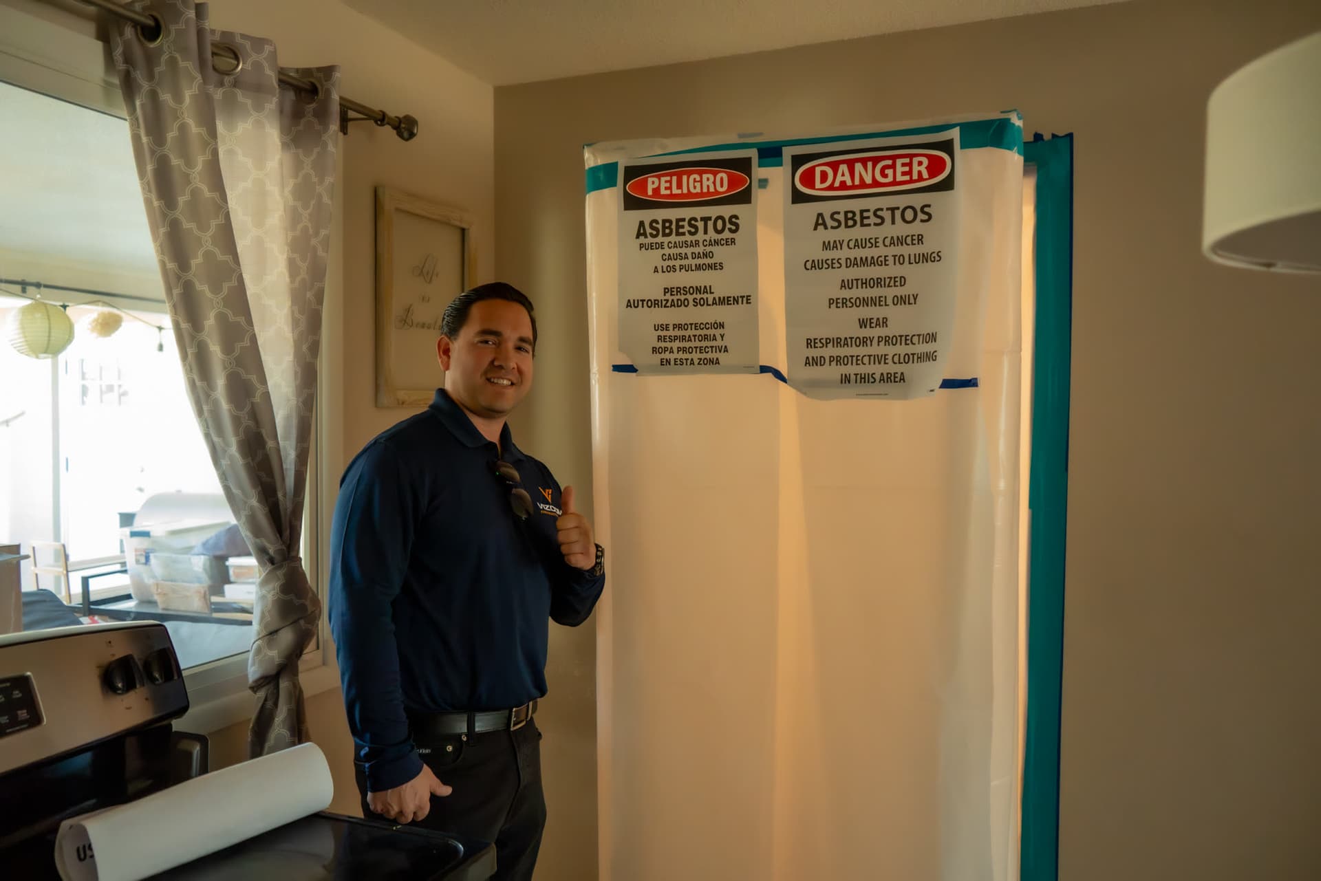 Omar Vizcarra standing beside a sealed plastic containment doorway inside a Phoenix kitchen, marked with asbestos danger signs in English and Spanish.