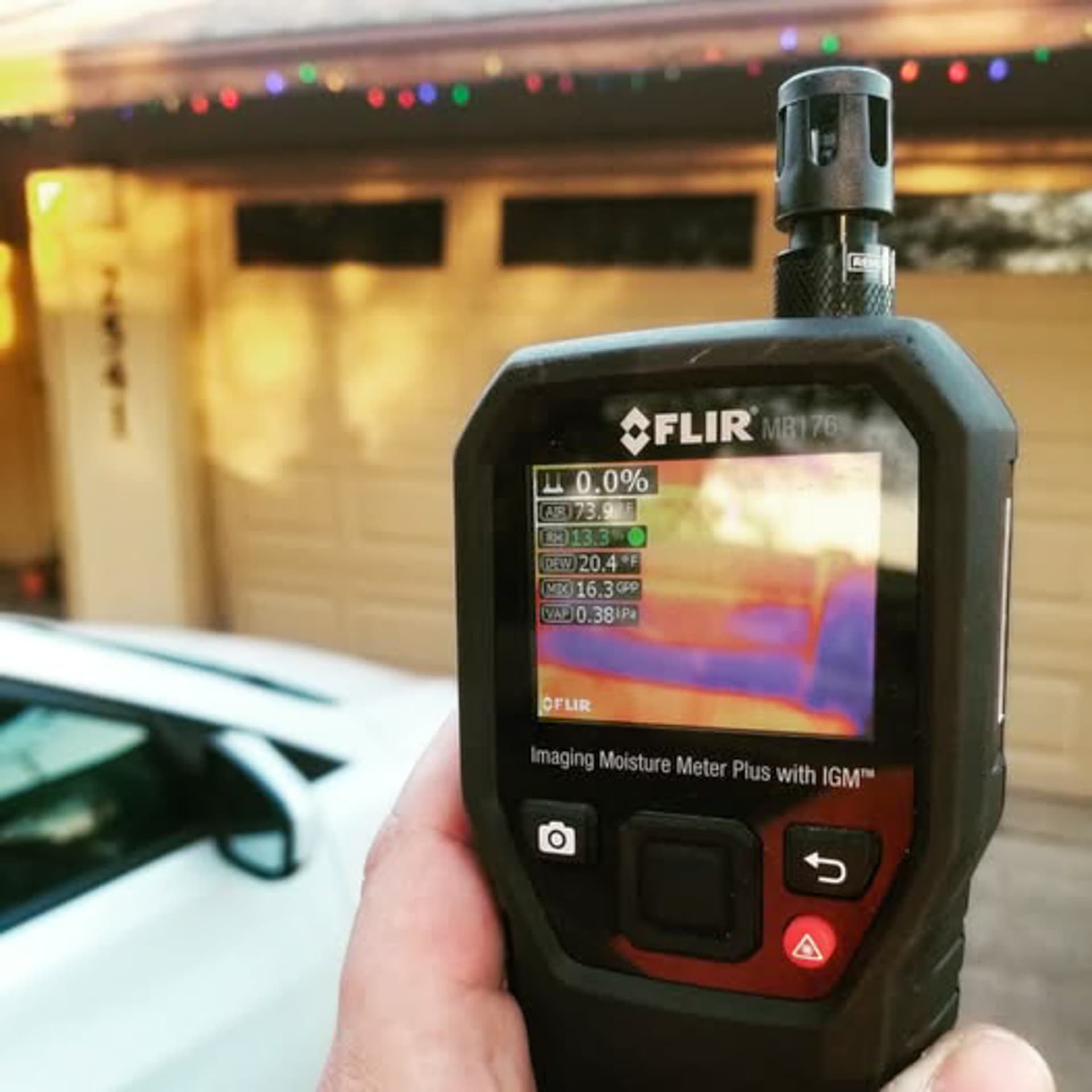 A thermal imaging moisture meter held up in front of a house, showing a cold band where water is sitting.